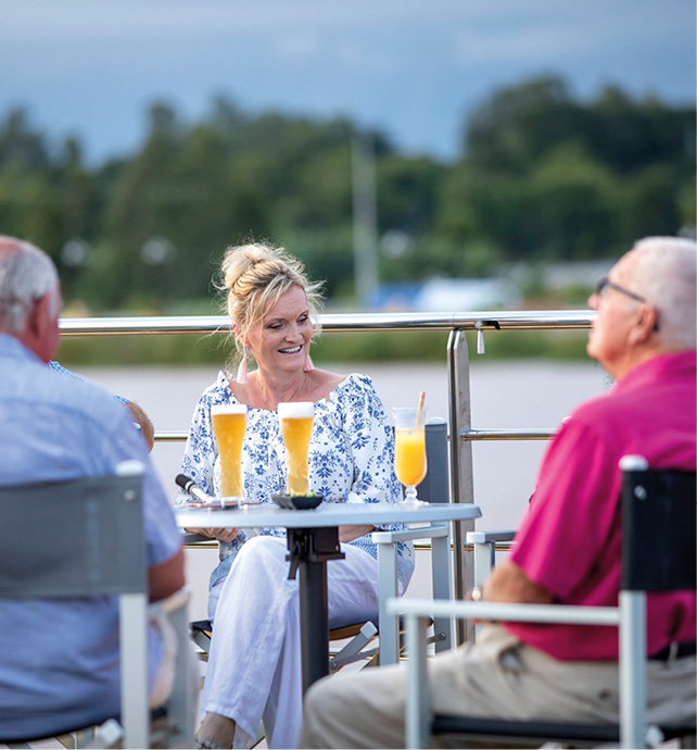 Five cruise guests enjoying beer and orange juice while enjoying the scenery found along the Mekong River, including the green trees