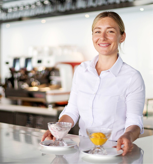 Smiling waitress placing two gelato servings on counter, with a coffee machine behind her, and a stack of plates to the side